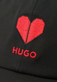 Black fabric with red embroidered broken heart shape and the word "HUGO" stitched below the heart.