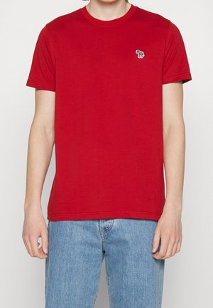 Mand iført ensfarvet rød kortærmet t-shirt med lille zebra-logo på brystet, kombineret med lyseblå denimjeans på hvid baggrund.