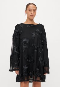 Melna kleita ar ziedu mežģīņu izšuvumu, garām caurspīdīgām piedurknēm, brīvu piegriezumu un laivu veida kakla izgriezumu, ar slāņotu faktūru apakšmalā.