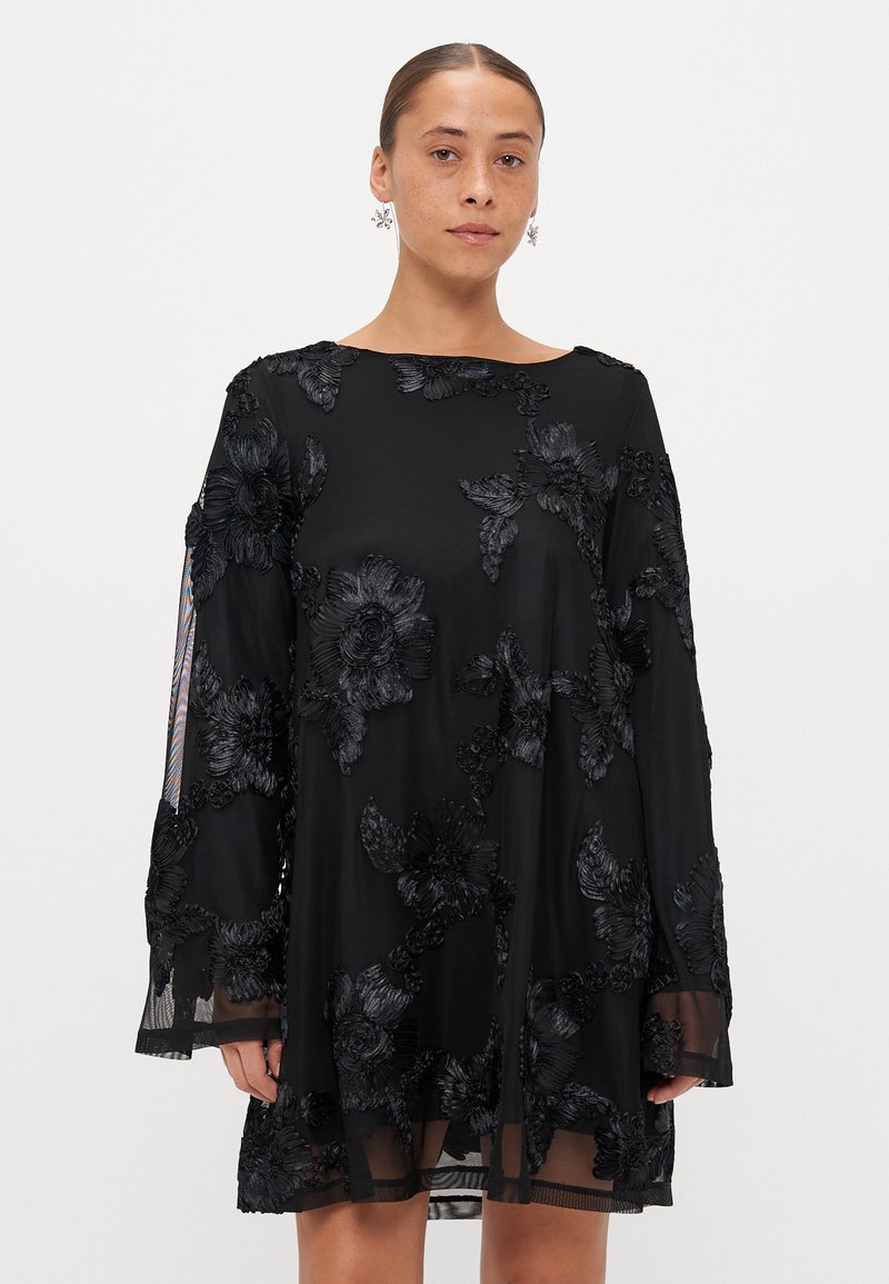 Melna kleita ar ziedu mežģīņu izšuvumu, garām caurspīdīgām piedurknēm, brīvu piegriezumu un laivu veida kakla izgriezumu, ar slāņotu faktūru apakšmalā.