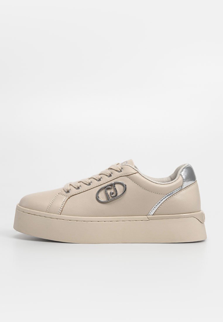 Zapatilla de cuero beige con acabado texturizado, puntera redonda, acentos plateados y suela de plataforma gruesa. Presenta un logotipo prominente en el costado.