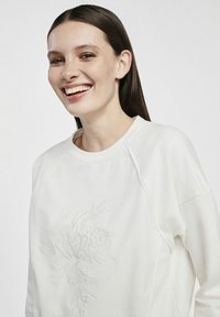 Kremowy sweatshirt z wypukłym kwiatowym wzorem, z okrągłym dekoltem i rękawami raglanowymi, wykonany z miękkiej tkaniny o gładkiej strukturze.