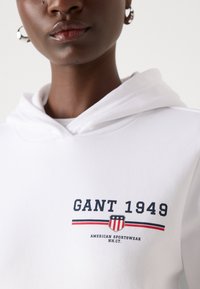 Bílá mikina s kapucí, která má na přední straně grafiku "GANT 1949", design štítu a červené a modré pruhy. Měkká tkanina s volným střihem.
