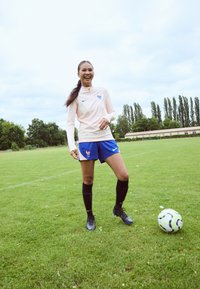 Sportininkas šviesiai rožinėje marškinėlių ilgomis rankovėmis su apykakle, mėlynose šortose, juodose iki kelių kojinėse ir juoduose sportiniuose bateliuose, stovi ant žalios žolės greta futbolo kamuolio.