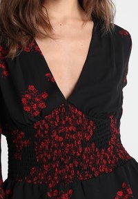 Schwarzes Kleid mit rotem Blumenmuster. Verfügt über einen tiefen V-Ausschnitt und eine geraffte Taille für eine figurbetonte Passform. Leichter Stoff.