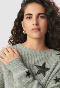 Jasnoszara sweter z dzianiny o teksturowym wykończeniu, z czarnymi skórzanymi aplikacjami w kształcie gwiazd oraz metalowymi koralikami zdobiącymi rękawy.