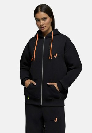 Femme portant un sweat à capuche noir zippé et un pantalon assorti avec des cordons orange et de petits détails de sangles, mains dans les poches du sweat, fond neutre.