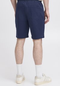 Tamsiai mėlyni šortai iš medvilnės, su elastiniu liemeniu, galine kišene ir tiesiu kirpimu, lygios tekstūros ir tvarkingai apdirgtomis kraštinėmis.
