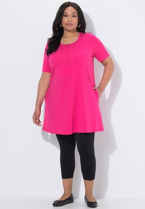 Kvinde iført en lyserød kortærmet tunika med lynlåsdetalje foran, sorte cropped leggings og sorte flade sko, stående med den ene hånd i lommen.