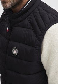 Svart boblevest med kviltet design, høy krage, rød og hvit glidelåsdrager, og sirkulært logo-patch på brystet. Ullkremgenser under.