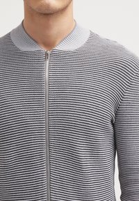 Gray and white striped zip-up cardigan with a ribbed collar, knitted texture, and metallic zipper detail. Features a fitted design.