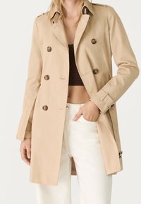 Femme portant un trench-coat beige sur un crop top marron et un pantalon blanc, avec une main dans la poche, sur fond uni.