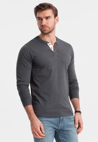 Man som bär en mörkgrå långärmad henley-tröja med träknappar och ljusblå jeans, stående mot en enkel vit bakgrund.