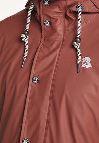 Vattentät röd jacka med en dragsnoddshuva med marinblå och vit rep, metalliska detaljer och en logotypapplikation på fram sidan.