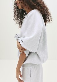 Frau mit lockigem Haar, die einen hellgrauen übergroßen Sweatshirt und passende Hose trägt, während sie den Ärmel einstellt, vor einem schlichten hellen Hintergrund.