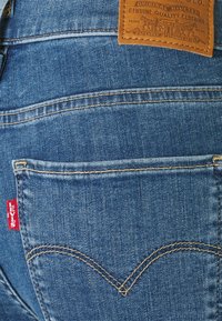 Vaqueros de denim azul con un parche de cuero marrón y etiqueta roja de Levi's. Presenta un diseño distintivo en los bolsillos traseros con un patrón de costura curvado.