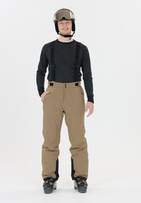 Beige Ski-Hosen mit verstellbaren schwarzen Trägern, ausgestattet mit zwei Fronttaschen und schwarzen Akzenten an den Knöcheln. Getragen mit einem grauen Langarmshirt.