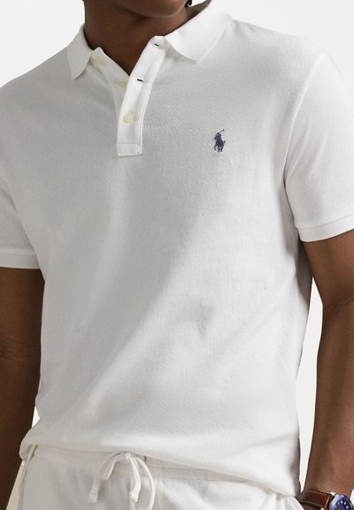 Hvit polo skjorte i en teksturert stoff, med knapplist med tre knapper, gule knapper og en kontrasterende blå logo på brystet.