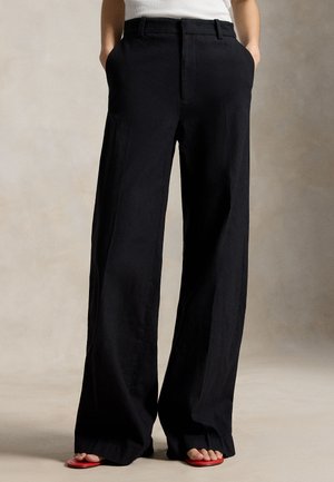 Čierne nohavice so širokými nohavicami a záhybmi, nosené s bielym topom a červenými sandálmi s otvorenou špičkou, ruky zasunuté vreckách.