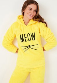 Set de pijama galben din fleece, cu glugă. Prezintă urechi de pisică, text imprimat "MEOW" și un design cu mustăți. Textură moale, croi casual.
