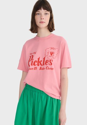 Young woman with short brown hair wearing pink T-shirt with red dog graphic and text "Pickles" paired with high-waisted green skirt.