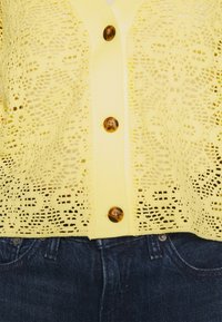 Geel kant cardigan met een knoopsluiting aan de voorkant, voorzien van schildpaddenknopen, gecombineerd met donkere denim jeans. Fijne details in het kantpatroon.