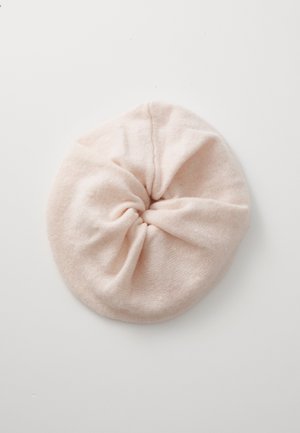 Miękka różowa dziergana beretka z centralnym marszczeniem, w okrągłym kształcie i gładkiej fakturze, leżąca na jednolitej białej powierzchni.