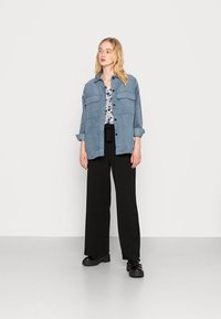 Camisa de denim azul-claro com dois bolsos no peito, usada sobre uma blusa branca de flores. Calças largas pretas e sapatos plataforma pretos completam o visual.