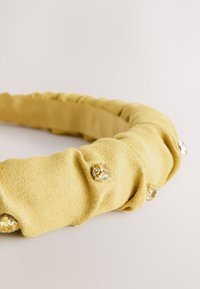 Yellow fabric headband with gathered texture, featuring metallic accents. Soft material with a smooth finish. Round shape, flexible design.