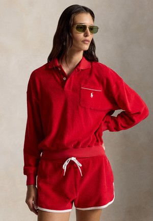 Kvinde iført rød langærmet polo med brystlomme og matchende røde shorts med hvide kanter, poserer med hånden på hoften, iført tonede solbriller.