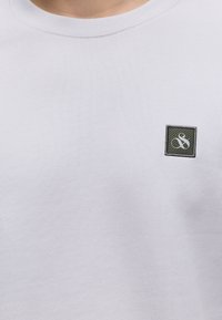 Λευκό μπλουζάκι με στρογγυλή λαιμόκοψη και μικρό σκούρο τετράγωνο λογότυπο με στυλιζαρισμένο λευκό αμперσάν στη αριστερή πλευρά του στήθους.