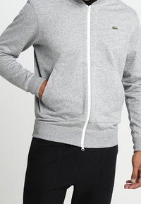 Šedá mikina na zip z měkké látky, se bílým zipem a malým zeleným logem na levé straně hrudi. Ukončeno žebrovanými manžetami.