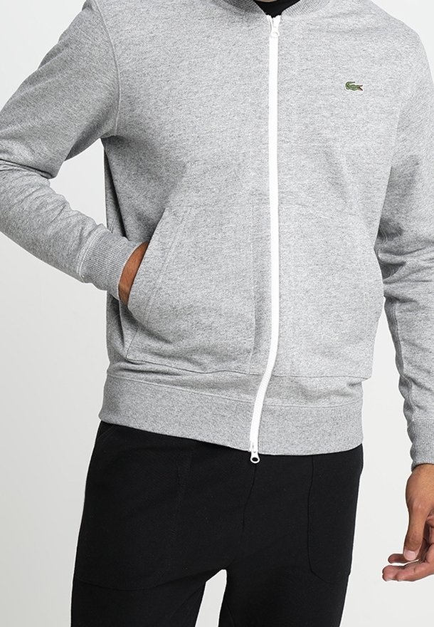 Šedá mikina na zip z měkké látky, se bílým zipem a malým zeleným logem na levé straně hrudi. Ukončeno žebrovanými manžetami.
