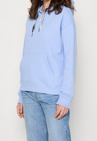 Person som bär en ljusblå hoodie med svarta och vita snören samt ljusblå jeans, stående mot en enfärgad bakgrund.