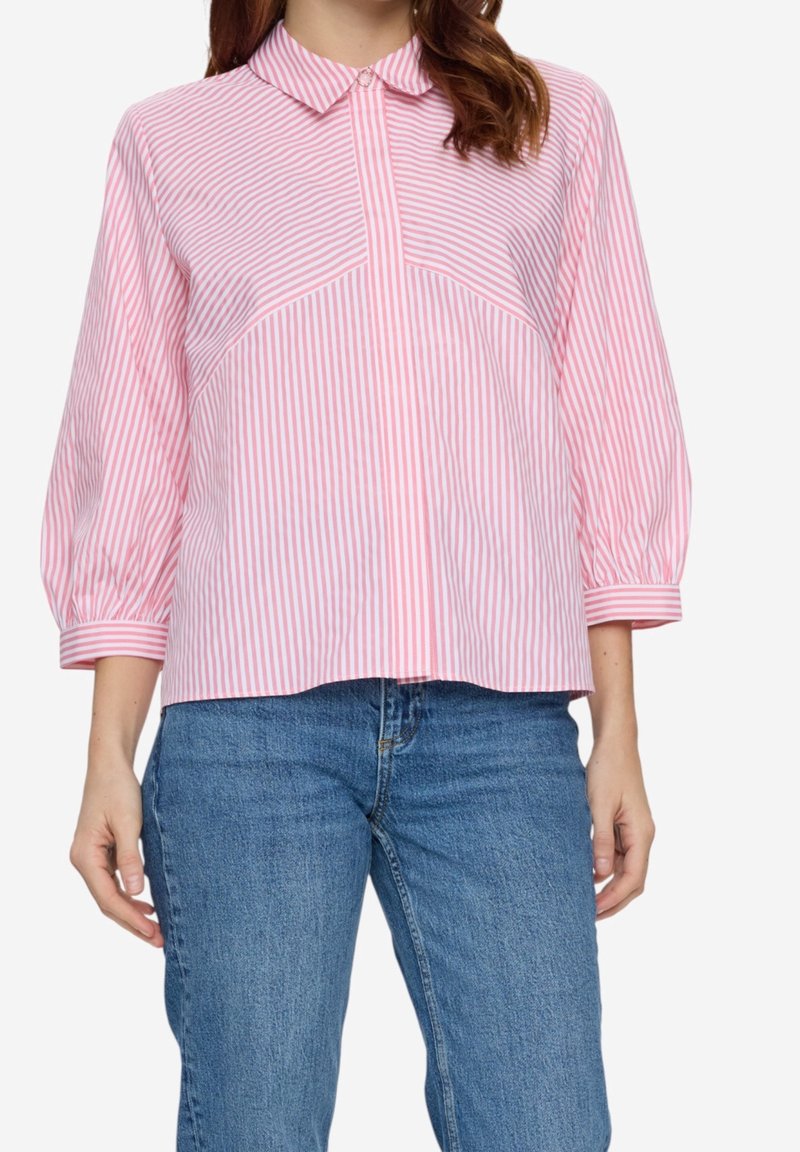 Femme portant un chemisier rayé verticalement rose et blanc avec des manches trois-quarts et un jean en denim bleu.