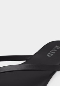 Κοντινό πλάνο ενός μαύρου σανδαλιού με λεπτές λουρίδες και επίπεδη σόλα, που εμφανίζει μέρος του τυπωμένου ονόματος της μάρκας στην εσωτερική σόλα.