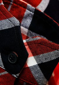 Camisa de franela de cuadros rojos y negros en primer plano, con finos patrones de cuadros, un botón negro en el cuello y una textura cepillada.