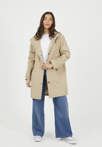 Beżowy wodoodporny płaszcz z kapturem, zamek błyskawiczny z przodu i zatrzaskami. Stylizowany z szerokimi niebieskimi dżinsami i białymi sneakersami. Gładka faktura.