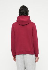 Burgundi värvi kapuutsiga sweatshirt, millel on vabalt kukkuv lõige, elastsed mansetid ja alumine äär; nähtav harjatud siseviimistlus. Kanda heledate hallide pükste peal.