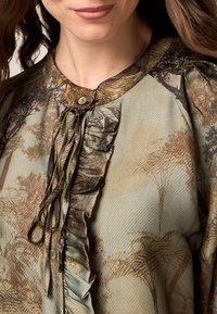 Bluzka z długim rękawem z miękkiego materiału, zielona z brązowym nadrukiem w drzewa, z okrągłym kołnierzykiem, falbaniastym przodem oraz zapięciem na guziki.