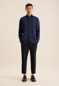 Marineblauw shirt met lange mouwen en knopen aan de voorkant, getailleerde pasvorm, gecombineerd met donkere broek en zwarte schoenen, tegen een neutrale achtergrond.