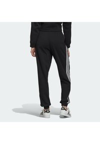 Person som bär svarta träningsbyxor med tre vita sidostråk och vita Adidas sneakers, stående mot en enfärgad bakgrund.