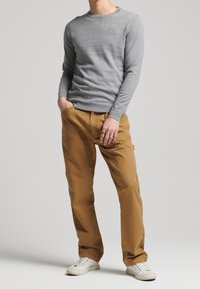 Pull gris clair à col rond, associé à un chino beige et des baskets blanches. Les détails notables comprennent un logo sur le pull et des poches sur le pantalon.