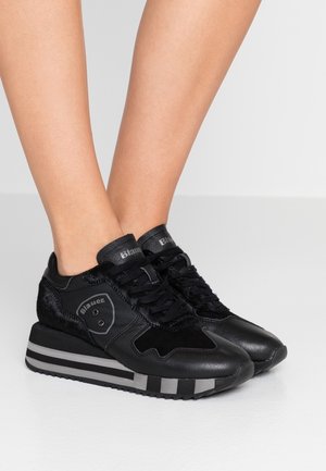 Nogi noszące czarne trampki Blauer z mieszanki skóry i zamszu, z grubymi paskowanymi podeszwami i sznurowanym fasonem, na jasnoszarej podłodze.