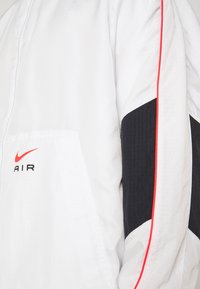Bílá větrovka s černým texturovaným panelem a červeným akcentním pruhem. Na přední straně je černé logo "AIR" pod Nike swoosh.