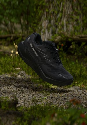 Chaussure de trail Salomon noire avec Gore-Tex, flottant au-dessus d'un sol rocheux dans un environnement forestier verdoyant.