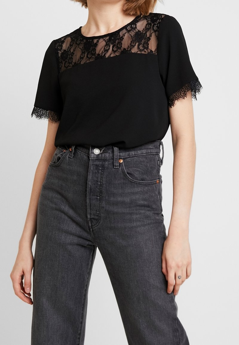 Femme portant un haut en dentelle noire à manches courtes et un jean taille haute gris foncé sur un fond uni.