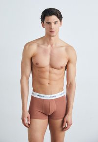 Barna pamut boxer rövidnadrág fehér derékpánttal, rajta a Lacoste logo. Sima textúra és testhez álló dizájn. A modell karjait az oldalán tartja.