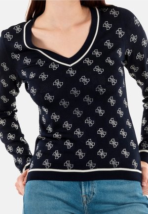 Femme portant un pull bleu marine à col en V avec des motifs floraux blancs et une bordure blanche, associé à un jean bleu clair, mains sur les hanches.