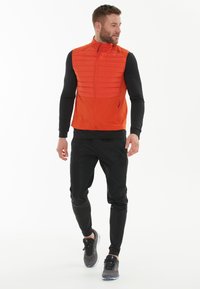 Orange vadderad väst med hög krage, dragkedja fram och två sidofickor, kombinerad med svarta träningsbyxor och grå sneakers.
